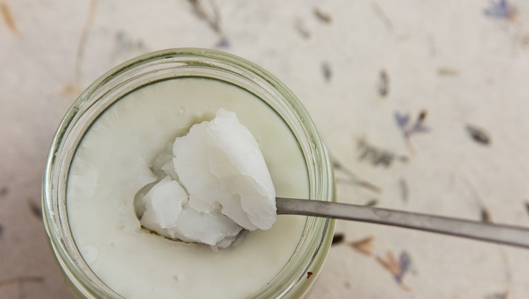 Coconut oil in a glass jar. Spoon with solidified oil. Out of focus decorative surface. High point of view. Light effect.